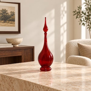 May include: A tall, translucent red glass decanter featuring a bulbous base, a slender elongated neck, and a pointed teardrop-shaped stopper. The vessel sits on a circular pedestal base, showcasing a smooth, reflective surface throughout its design.