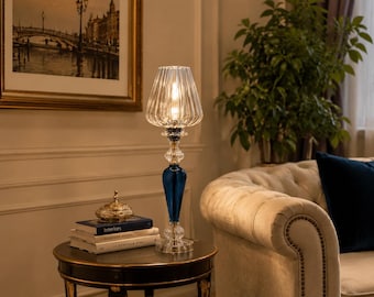 May include: A tall table lamp featuring a clear, ribbed glass shade and a slender, multi-sectioned base. The base includes a deep blue, teardrop-shaped glass element accented by clear, textured glass segments at the top and bottom, resting on a circular base.