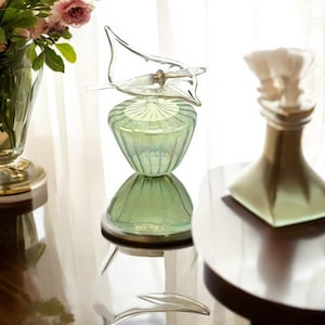 Può includere: Una bottiglia di vetro verde con un tappo di vetro trasparente a forma di uccello. La bottiglia è appoggiata su un tavolo di legno con uno sfondo di stoffa bianca.