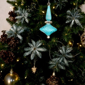 Puede incluir: Un adorno de Navidad de vidrio azul con un dise&ntilde;o de remolino cuelga de un &aacute;rbol de Navidad verde. El adorno tiene forma de estrella y tiene una punta plateada.