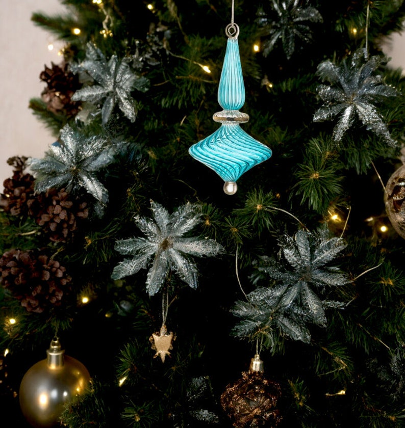 Puede incluir: Un adorno de Navidad de vidrio azul con un dise&ntilde;o de remolino cuelga de un &aacute;rbol de Navidad verde. El adorno tiene forma de estrella y tiene una punta plateada.