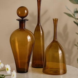 Puede incluir: Tres botellas decorativas de vidrio ámbar de varias formas. Una botella tiene un tapón redondo, otra es alta y delgada, y la tercera tiene una base acampanada. Las botellas están sobre una mesa de mármol blanco.