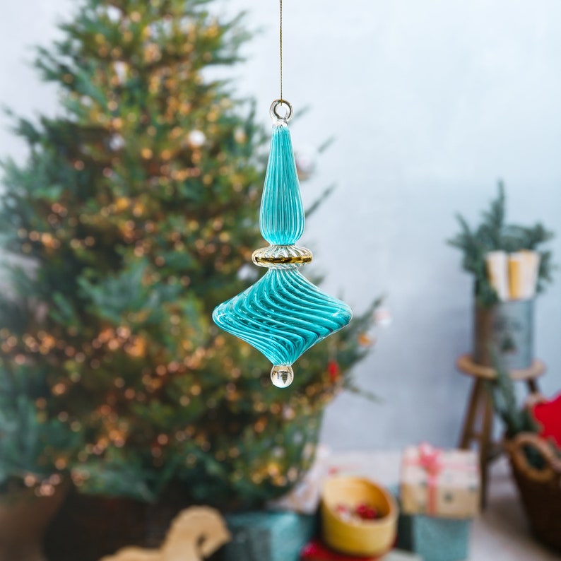 Puede incluir: Un adorno navide&ntilde;o de vidrio azul turquesa con ribete dorado y dise&ntilde;o en espiral. El adorno est&aacute; colgado de una cuerda.