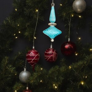 Puede incluir: Un adorno de Navidad de vidrio azul con un dise&ntilde;o de espiral cuelga de una cadena de luces en un &aacute;rbol de Navidad verde. Tambi&eacute;n hay adornos rojos y plateados en el &aacute;rbol.