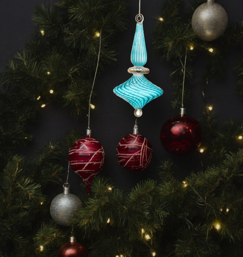 Puede incluir: Un adorno de Navidad de vidrio azul con un dise&ntilde;o de espiral cuelga de una cadena de luces en un &aacute;rbol de Navidad verde. Tambi&eacute;n hay adornos rojos y plateados en el &aacute;rbol.
