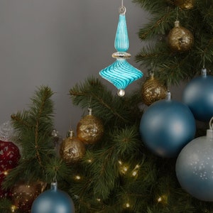 Puede incluir: Un adorno de Navidad de vidrio azul con un dise&ntilde;o de espiral cuelga de un &aacute;rbol de Navidad con otros adornos, incluidos adornos dorados y azules.