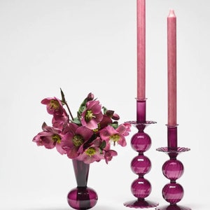 May include: Three purple glass candle holders with pink candles. The candle holders are shaped like a ball with a scalloped base. The candles are tall and thin. A purple glass vase with pink flowers is in the background.
