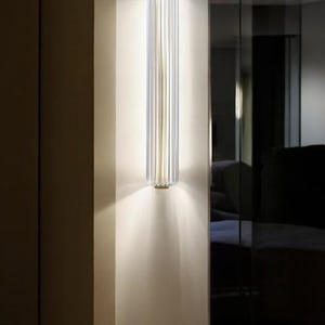 May include: A modern wall sconce with a clear glass shade and a gold metal frame. The sconce is mounted on a white wall and casts a warm glow.