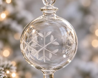 Puede incluir: Un adorno de cristal transparente de forma esférica con un delicado patrón grabado en blanco que representa hojas o un copo de nieve. El adorno tiene un pequeño lazo en la parte superior para colgarlo y una base redondeada en forma de gota.