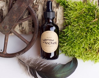 May include: A brown glass bottle with a dropper top and a label that reads "Wildcrafted Yarrow Tincture Good Green Gardens Gluten Free". The bottle is sitting on a piece of birch bark with green moss and a brown feather.