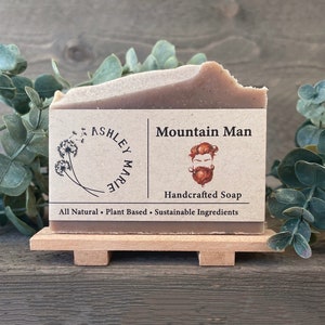 May include: A brown bar of soap with a label that reads "Mountain Man Handcrafted Soap" and "All Natural - Plant Based - Sustainable Ingredients". The label also features a cartoon illustration of a man with a beard.