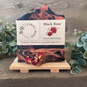 May include: A bar of handmade soap with a black and red marbled design. The soap is decorated with dried rose petals and has a label that reads "Black Rose Handcrafted Soap All Natural Plant Based Sustainable Ingredients".