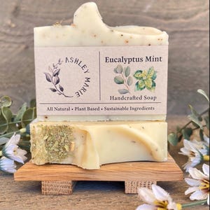 May include: Two bars of handcrafted Eucalyptus Mint soap, one atop the other, on a small wooden soap dish. The top bar has a curved shape and a label that reads "Ashley Marie" and "Eucalyptus Mint, Handcrafted Soap." The soap is light green with visible herbs.