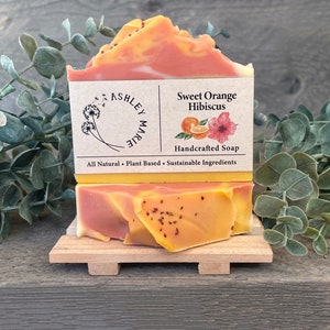 Puede incluir: Una pastilla de jabón artesanal con aroma a naranja dulce e hibisco. El jabón es de color naranja y rosa vibrante con un patrón de remolino. La etiqueta dice "Jabón artesanal de naranja dulce e hibisco" y "Todo natural - A base de plantas - Ingredientes sostenibles".