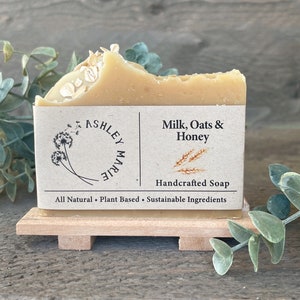 May include: A bar of handcrafted soap with a label that reads "Milk, Oats & Honey" and "Handcrafted Soap". The label also says "Ashley Marie" and "All Natural - Plant Based - Sustainable Ingredients".