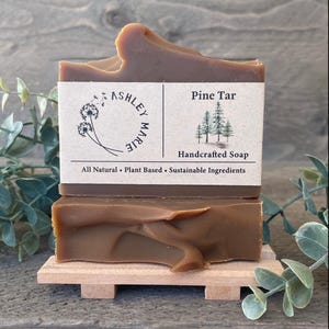 Può includere: Due saponette artigianali al catrame di pino sono esposte su una piccola piattaforma di legno. Il sapone è di colore marrone medio con una superficie strutturata. Un'etichetta sul sapone recita "Pine Tar Handcrafted Soap" e "All Natural • Plant Based • Sustainable Ingredients."