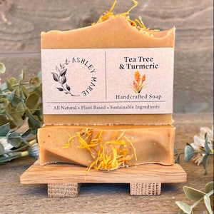 Op de afbeelding: Twee handgemaakte zeepblokken met de tekst "Tea Tree & Turmeric" en "Ashley Marie" op een houten zeepbakje. De zeep is lichtbruin met gele accenten en gedroogde bloemblaadjes. Het zeepbakje is gemaakt van lichtbruin hout.