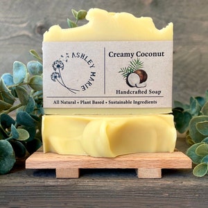 May include: A yellow bar of handmade soap on a wooden soap dish. The soap is labeled "Creamy Coconut" and "Ashley Marie". The label also says "All Natural - Plant Based - Sustainable Ingredients".