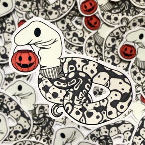 Puede incluir: Una pegatina con una serpiente de dibujos animados con cabeza color crema y cuerpo con estampado en blanco y negro. La serpiente sostiene una linterna de calabaza y está rodeada de pegatinas similares, creando un tema de Halloween.