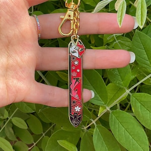 May include: A gold keychain with a red and white enamel charm featuring a stylized fox design. The charm is attached to a gold split ring and a gold chain.