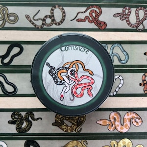 Op de afbeelding: Een ronde sticker met een groene rand en een witte achtergrond. De sticker toont een tekening van drie in elkaar verstrengelde maisslangen, met de tekst "cornsnake" in zwart bovenaan.