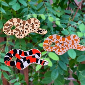 Può includere: Tre spille a forma di serpente in smalto colorato. Una è beige con macchie marroni, un'altra arancione con macchie bianche e la terza rossa, nera e bianca. Le spille sono poste su uno sfondo di foglie verdi.