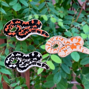 Op de afbeelding: Drie emaille pins in de vorm van slangen. Eén pin is zwart en wit, een andere is oranje en zwart, en de derde is oranje en crème. De pins hebben goudkleurige contouren en zijn geplaatst tegen een achtergrond van groene bladeren.