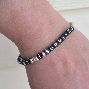 Pu&ograve; includere: Braccialetto di perline nere con scritta bianca "I SURVIVED" sul polso di una persona.