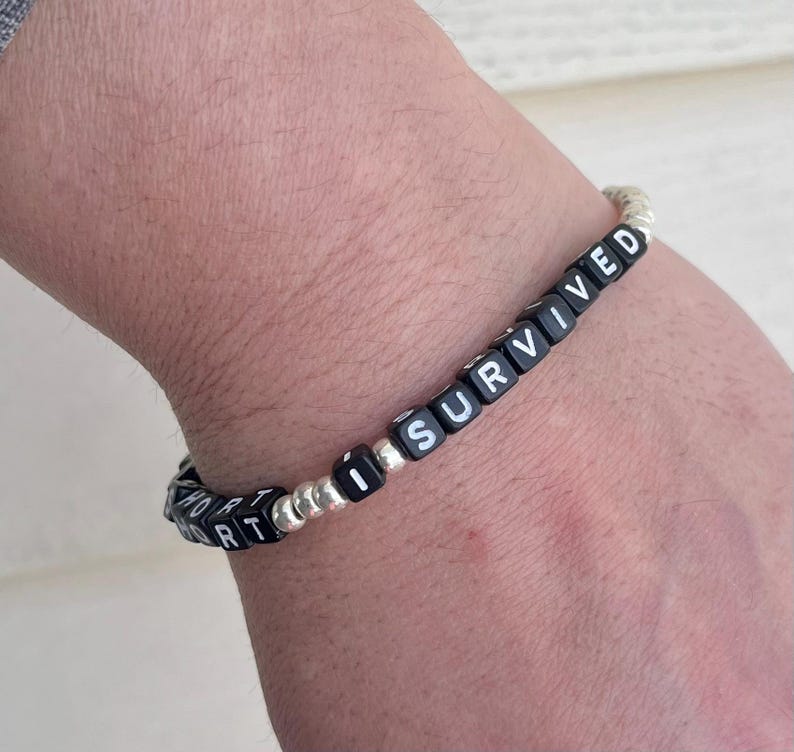 Pu&ograve; includere: Braccialetto di perline nere con scritta bianca "I SURVIVED" sul polso di una persona.