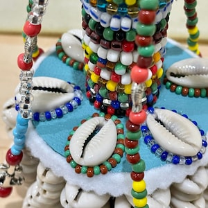 Może przedstawiać: Biała ceramiczna podstawa z kolorową kolumną koralików i białą obwódką z muszli. Podstawa jest ozdobiona różnymi kolorowymi koralikami, w tym czerwonymi, niebieskimi, zielonymi, żółtymi i białymi. Podstawa jest również ozdobiona białymi muszlami.