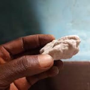 May include: A hand holding a small, irregularly shaped, light-coloured rock. The rock has a rough, textured surface. The background is a blurred mix of blue and brown.