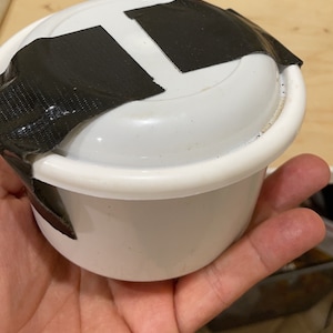 May include: A white plastic container with a black tape seal on the lid.