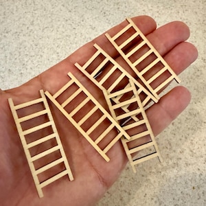 Puede incluir: Un conjunto de pequeñas escaleras de madera sin terminar. Cada escalera tiene peldaños espaciados uniformemente y un diseño sencillo. Las escaleras se sostienen en una mano, mostrando su tamaño y detalle. Ideales para manualidades o proyectos en miniatura.