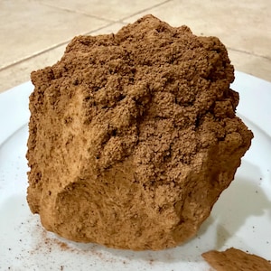 Puede incluir: Un objeto grande, texturizado y marrón, posiblemente un postre, se encuentra sobre un plato blanco. El objeto tiene una superficie rugosa y desmenuzable y un color marrón claro. Se ha roto un trozo pequeño.