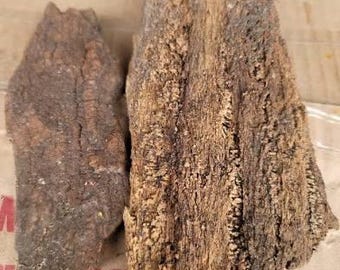 May include: Two pieces of weathered wood with a rough, textured surface. One piece is a dark brown color, while the other is a lighter tan with darker brown accents. The wood appears to be natural and aged.