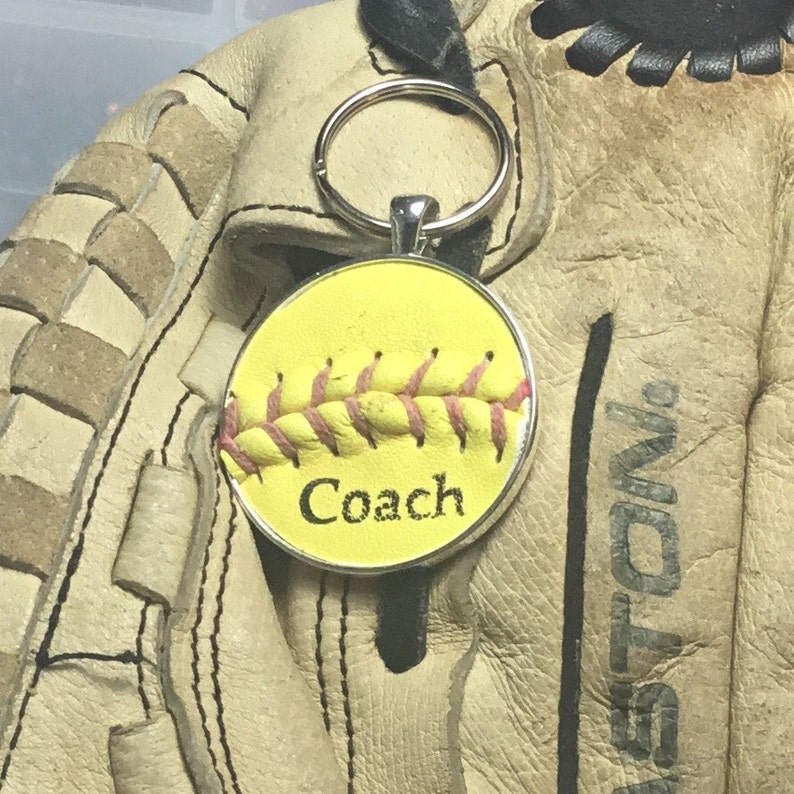 May include: Yellow softball keychain with pink stitching and the word "Coach" on a silver background. The keychain is attached to a silver key ring.