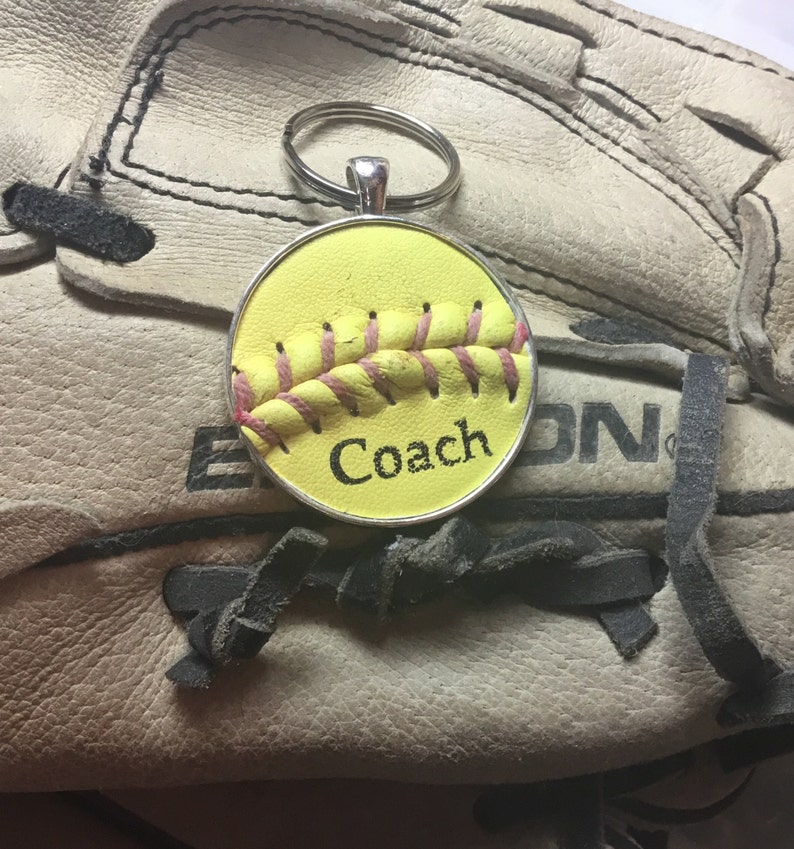 May include: A yellow softball keychain with pink stitching and the word "Coach" on it. The keychain is sitting on a brown leather softball glove.