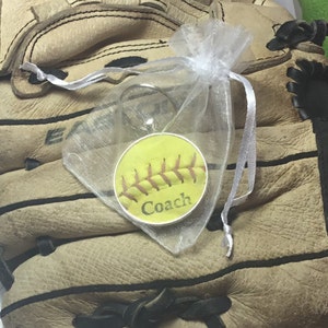 May include: A yellow softball keychain with red stitching and the word "Coach" printed on it. The keychain is in a white organza bag and is sitting on a brown leather baseball glove.