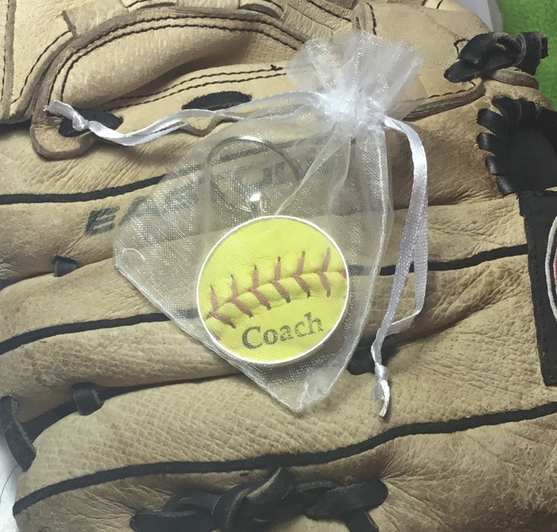 May include: A yellow softball keychain with red stitching and the word "Coach" printed on it. The keychain is in a white organza bag and is sitting on a brown leather baseball glove.