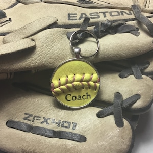 May include: A yellow softball keychain with pink stitching and the word "Coach" on it. The keychain is attached to a silver key ring and is sitting on top of a brown leather baseball glove.