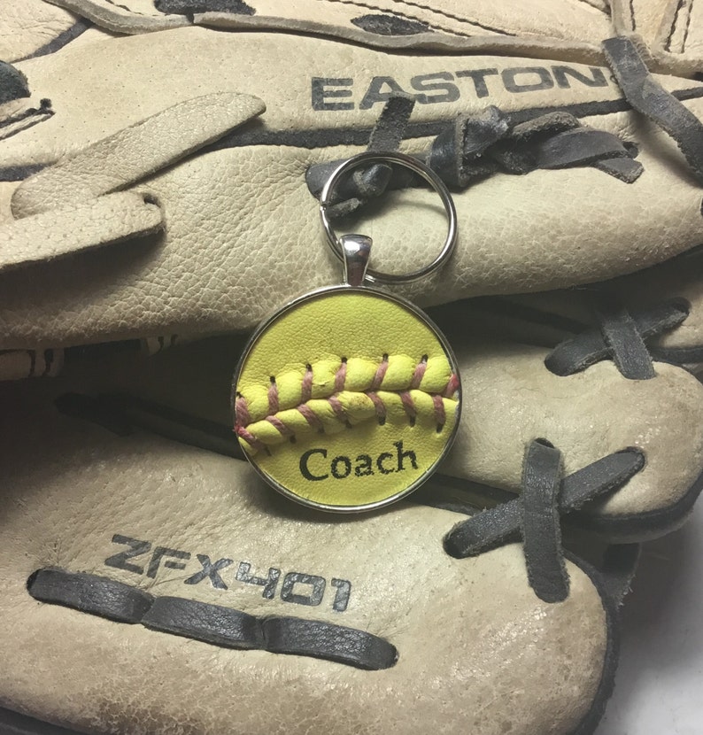 May include: A yellow softball keychain with pink stitching and the word "Coach" on it. The keychain is attached to a silver key ring and is sitting on top of a brown leather baseball glove.