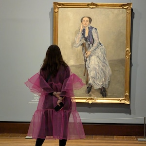 May include: A large oil painting in a gold ornate frame depicting a seated figure wearing a blue and white striped dress with a dark blue shawl. The figure rests their chin on one hand while sitting on a wooden chair against a neutral background.