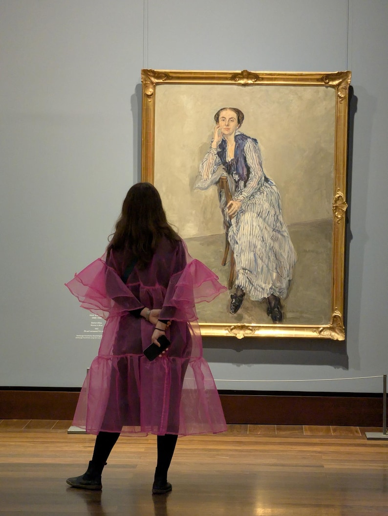 May include: A large oil painting in a gold ornate frame depicting a seated figure wearing a blue and white striped dress with a dark blue shawl. The figure rests their chin on one hand while sitting on a wooden chair against a neutral background.