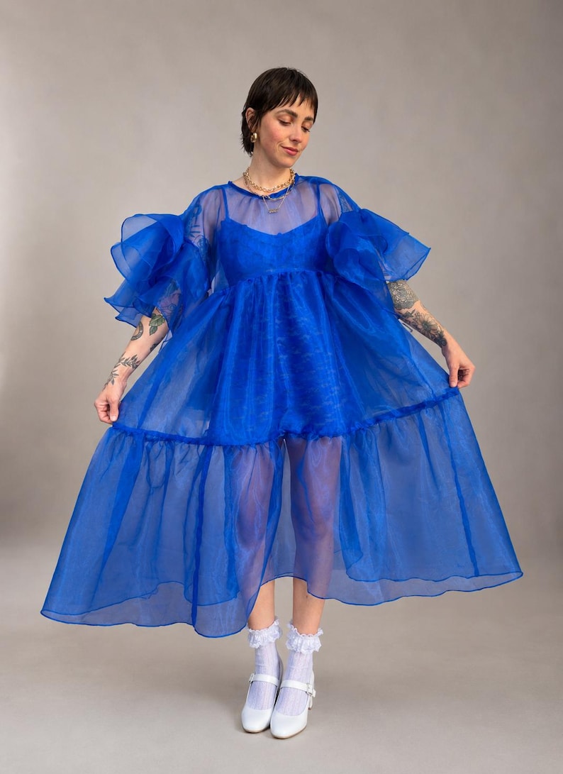May include: A vibrant royal blue sheer organza dress featuring voluminous ruffled sleeves, a round neckline, and a tiered skirt silhouette. The garment is worn over a matching blue camisole, paired with white ruffled ankle socks and white shoes.