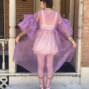 May include: A sheer lavender organza duster featuring voluminous tiered ruffled sleeves and a high-low hemline. The garment is worn over a white lace slip dress and paired with metallic pink cowboy boots, creating a layered look with visible texture.