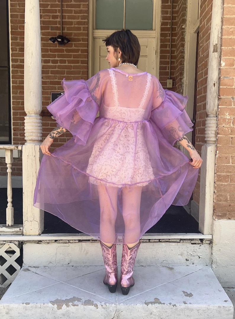 May include: A sheer lavender organza duster featuring voluminous tiered ruffled sleeves and a high-low hemline. The garment is worn over a white lace slip dress and paired with metallic pink cowboy boots, creating a layered look with visible texture.