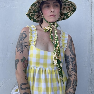 Op de afbeelding: Een vrouw draagt een groene en witte zonnehoed met bloemenprint en een bijpassende sjaal om de nek geknoopt. Ze draagt een geel-witte gingham jurk met ruches bandjes. De hoed heeft een brede rand.