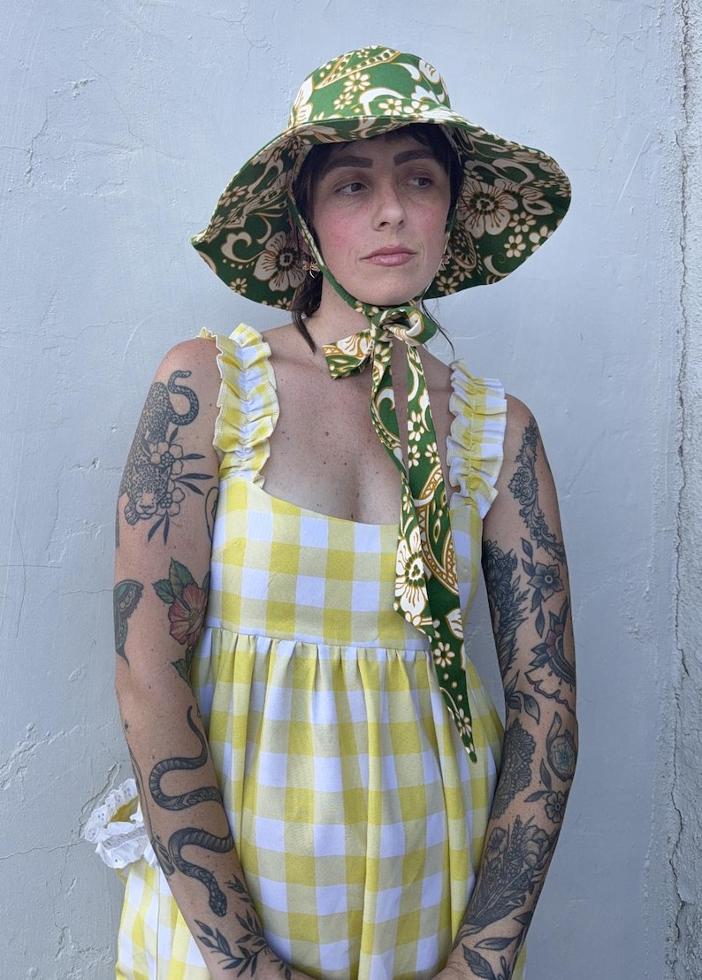 Op de afbeelding: Een vrouw draagt een groene en witte zonnehoed met bloemenprint en een bijpassende sjaal om de nek geknoopt. Ze draagt een geel-witte gingham jurk met ruches bandjes. De hoed heeft een brede rand.