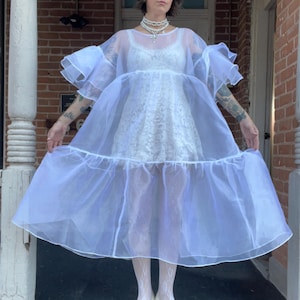 May include: A white sheer organza dress with voluminous ruffled sleeves and a tiered skirt, worn over a white lace mini dress. The ensemble is paired with white lace tights and white low-heeled shoes, accessorized with a layered pearl necklace.