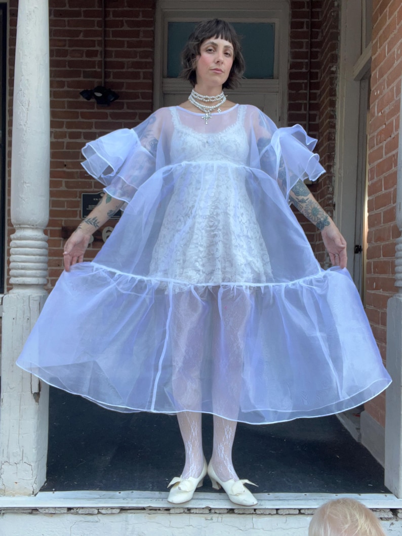 May include: A white sheer organza dress with voluminous ruffled sleeves and a tiered skirt, worn over a white lace mini dress. The ensemble is paired with white lace tights and white low-heeled shoes, accessorized with a layered pearl necklace.